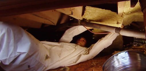白いジャンプスーツを着た男性が、家の下の天井裏の根太の間に断熱材を入れている様子。
