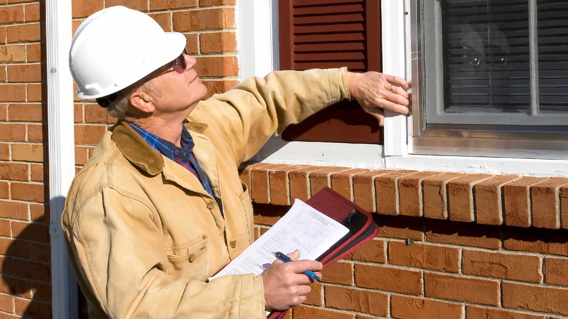 Home inspector checking windows on the outside of home and making notes on his clipboard.