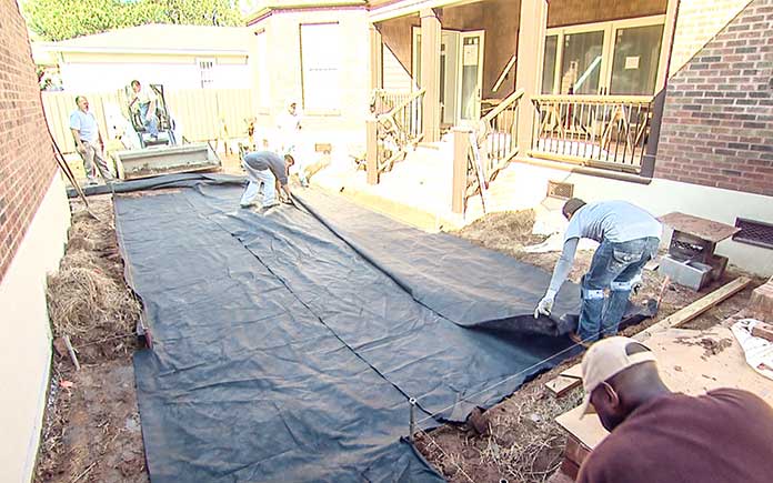 A team of workers lays landscape fabric over the excavation site of a paver patio