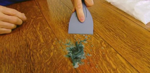 Using a plastic putty knife to remove hardened candle wax from a dining room table.