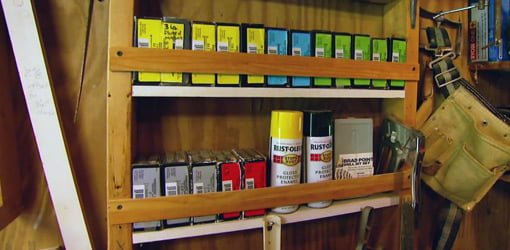 Shelves in open stud wall in a workshop filled with nail and screw boxes, spray paint cans, and hand tools..