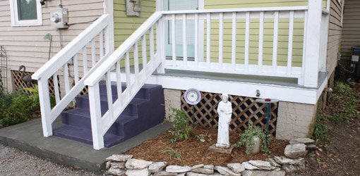 Wood porch with white railings, gray flooring, plumb steps, and yellow-green siding.