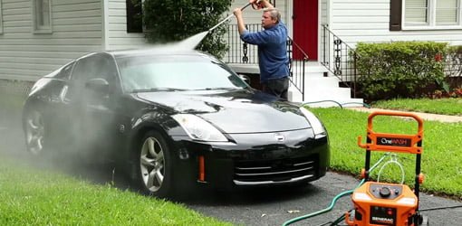 Man using a pressure washer to clean a black sports car in driveway.