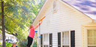 Woman pressure washes a home's wood siding as another woman holds the ladder that she is standing on.