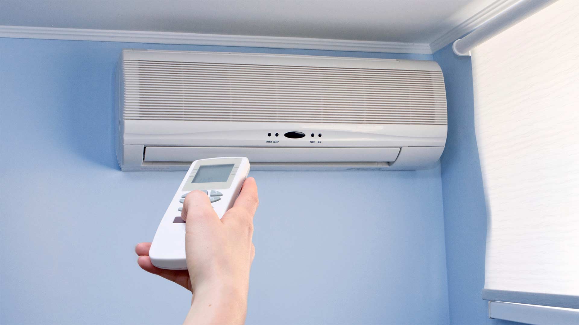 Person aiming remote control toward ductless mini-split air conditioner installed on a wall just below the ceiling.