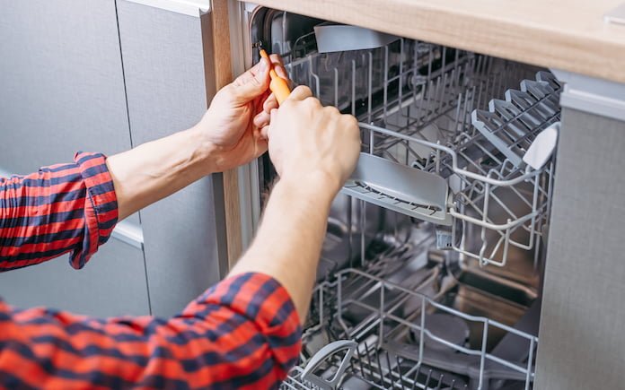 Man repairing dishwasher. Male hand with screwdriver installs kitchen appliances, close up