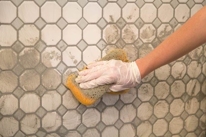 Gloved hand wipes grout off tile backsplash with a damp sponge