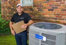 Friendly American Standard HVAC technician, who is wearing a cap, smiles, holds a clipboard, and stands beside a condenser unit