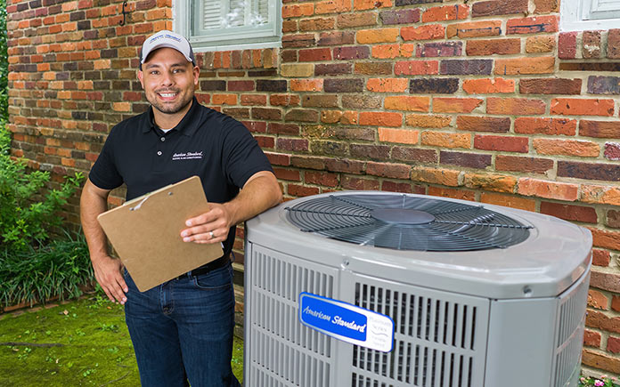 Friendly American Standard HVAC technician, who is wearing a cap, smiles, holds a clipboard, and stands beside a condenser unit