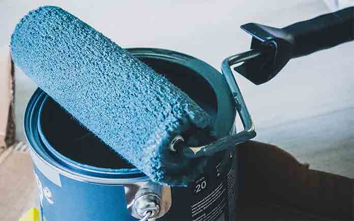 Paint roller with blue paint on it, resting on an open paint bucket