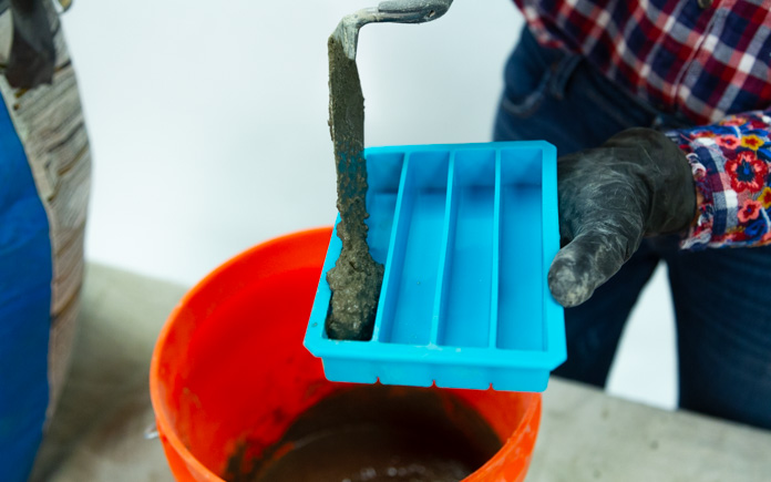 A gloved hand uses a masonry trowel to pour concrete mix into an ice tray