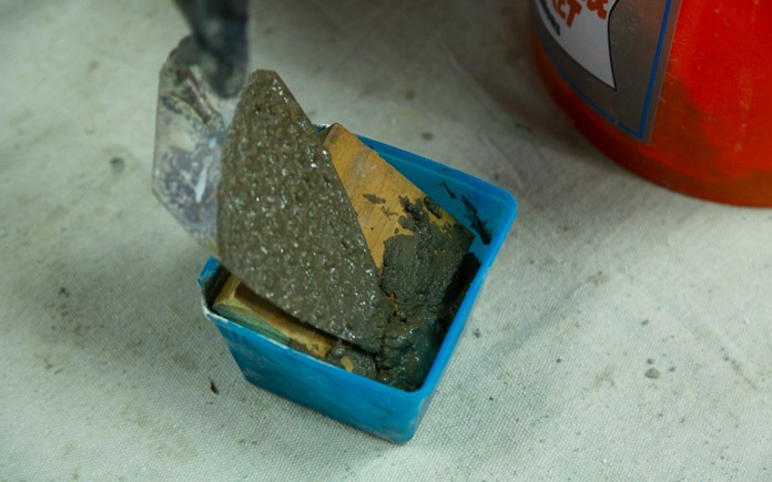A gloved hand uses a masonry trowel to lay a second pour of concrete mix on top of a wood cutout