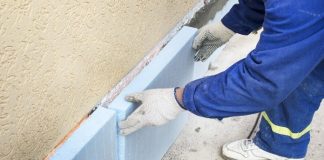 A person wearing a blue workmen suit, white gloves, and white sneakers repairs the foundation of a home.