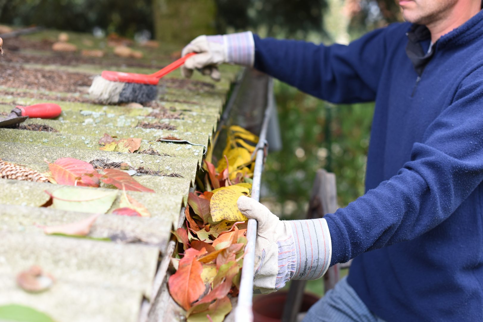 A person cleans out the gutters on the roof of a home while wearing white gloves and using a red brush.