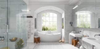 Large white updated bathroom with white and light grey tiled floors and ceilings, a walk in glass shower, white bathtub and 2 white sinks, with a window over the tub and trees in the background, simple decoration pieces