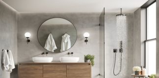A newly remodeled bathroom with grey walls, a wood vanity, rainfall walk in glass shower, white towels and robes, wound mirror over vanity, white sinks with black finishings