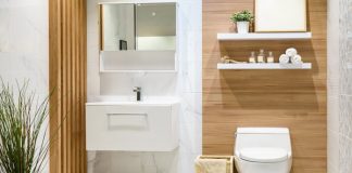 A newly remodeled bathroom with a white toilet, sink, shelves, and tiled walls. Wood finishings on the wall, and a wood separator, a mirror on the wall and a plant on the floor