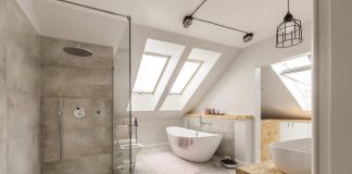 An updated bathroom with white walls, glass walk in shower, white toilet and bathtub, roof windows, minimalistic steel lighting fixtures, light brown counter tops