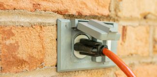 Power cord plugged into outdoor electrical wall socket. Dual covered electricity power outlets on brick wall. Orange extension cord plug inserted into exterior wall jack.