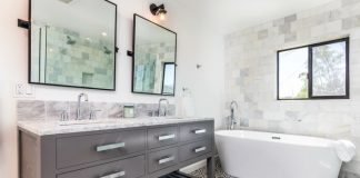 An updated bright modern bathroom, with a window over a white stand-alone tub, a sink with dark grey cabinets and light grey marbled countertops, with a black and white pattern tiled floor, and two vanity mirrors