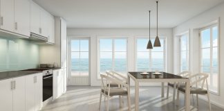 A beach house kitchen with white cabinets, stainless steel kitchen appliances, a table and chair with two light fixtures above, and panoramic windows with a blue ocean background