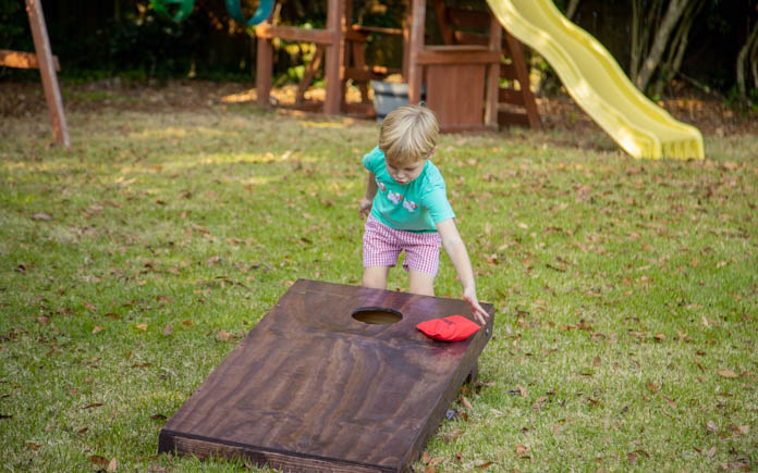 Child grabbing a cornhole bag from the top of a cornhole board game in a backyard.