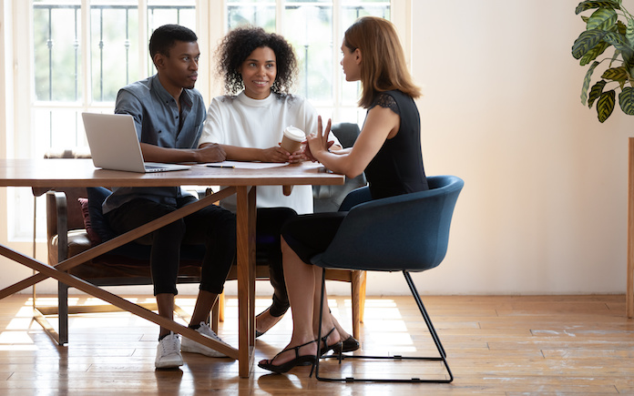 Young African American husband and wife sit at desk talk with female real estate agent discuss buying house together, biracial couple meet with designer architect or broker consult in modern office