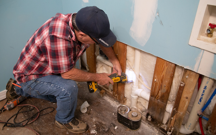 A plumber uses an oscillating saw on a white PVC pipe inside drywall