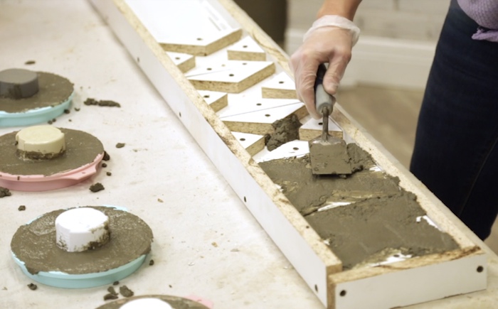 O-shaped silicone cake pans with concrete mix inside them and a trowel spreading concrete over a melamine form with x shapes 