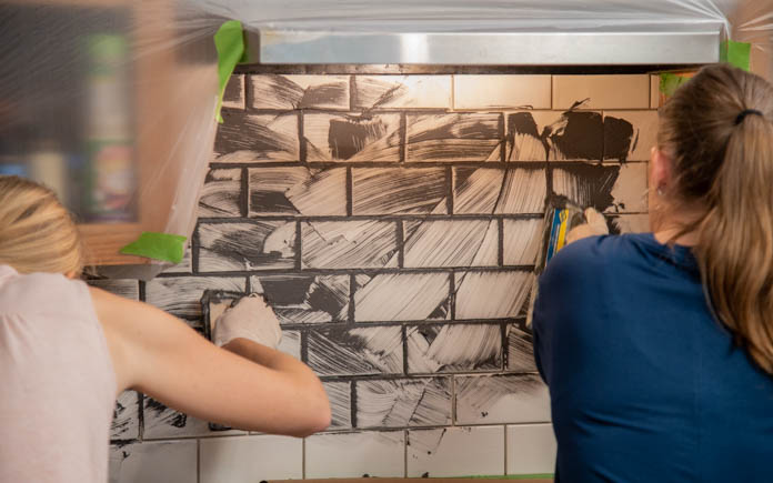 Two woman spread grout on a subway tile backsplash behind a stove