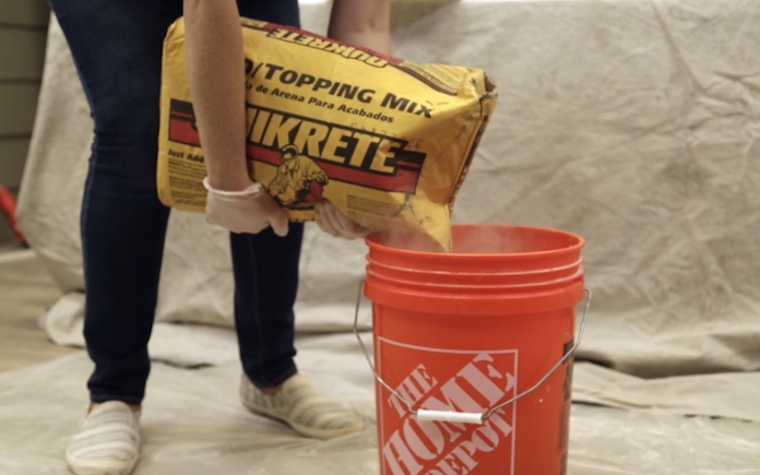 Gehandschoende handen die Quikrete Sand/Topping Mix in een oranje emmer van Home Depot gieten