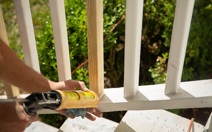 Hands use a caulk gun to apply caulk around a new wood spindle on a front porch handrail