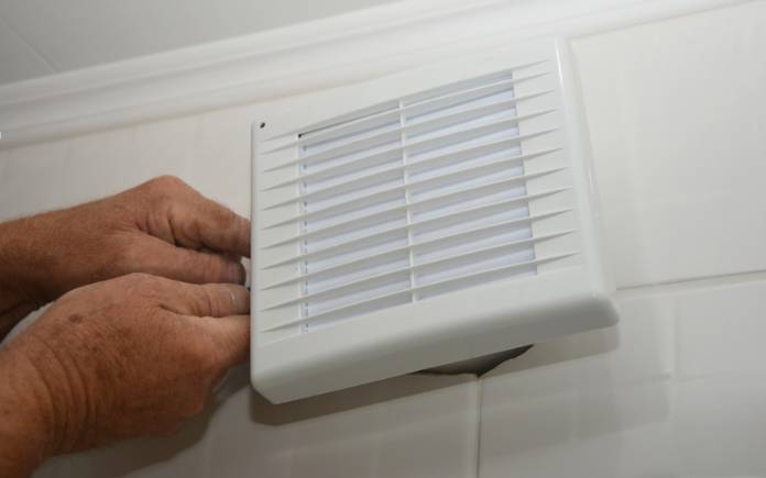 Hands installing a wall-mount bathroom exhaust fan on a tile wall